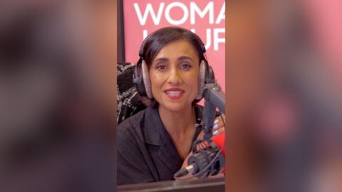 A woman with short, dark hair sits in front of the microphone and speaks to the camera