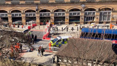 A large, historic-looking railway station building made of yellow-brown brick. The architecture features a row of tall, arched windows along the façade, each framed by stone pillars and decorative detailing.