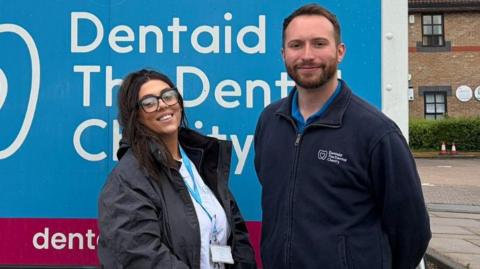 Christina, a young woman with glasses and brown hair, wearing a lilac blue charity-branded shirt. She is stood next to Ryan, a young man with short hair and a short beard, wearing a navy blue charity-branded jacket. They are stood in front of a van, which we can see is branded with the "Dentaid" logo.