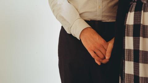 The middle portion of a woman standing up wearing trousers and a shirt. Her right hand appears to be holding another person's hand in front of her crotch, although the other person is hidden behind some patterned material. 