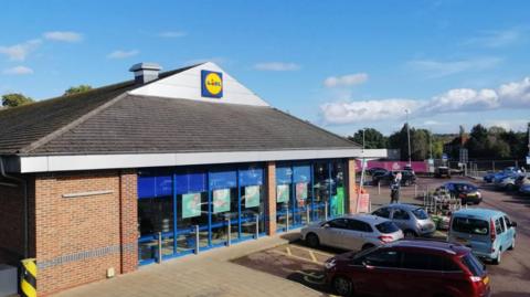 Lidl car park in Stourport. Cars are parked outside the supermarket with the Lidl company logo. A person is is outside the store. There is a blue sky.