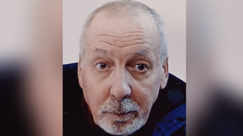 Portrait shot of Harry Murphy, who has short white hair and a grey beard. He is looking at the camera. The sides of the image are blurred.