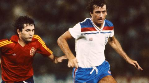 Trevor Francis is wearing a white Admiral manufactured England shirt and is being chased by a Spanish player wearing a red Adidas designed shirt. 