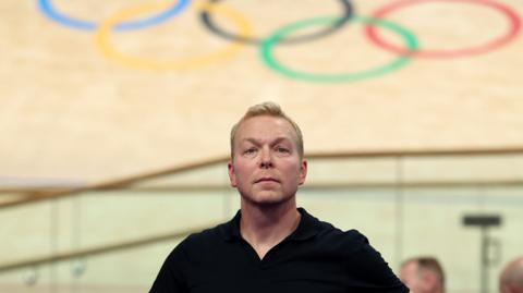 Sir Chris Hoyle, looking calm in a black top, stands below the Olympic rings