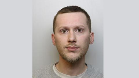 A police custody mugshot of Tony Heath. He has short dark hair and a small amount of facial hair. He is looking directly at the camera with a blank expression and is wearing a grey sweatshirt
