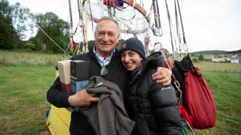 A middle aged man wearing a jumper and shirt has his arm round a young woman wearing a coat and beanie hat. They are stood in a field in front of a balloon.