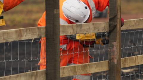 A person wearing orange high-viz and a white hard helmet is bending over holding a yellow power tool. They are working on a wooden fence. The ground is charred.