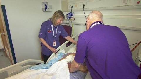 Two volunteers in purple t-shirts on either side of hospital bed of a patient whose face is obscured by the volunteer on the right