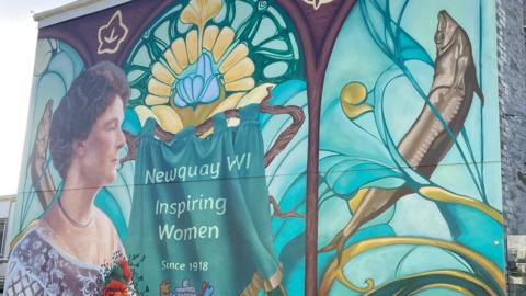 The mural shows Lady Molesworth-St Aubyn in profile wearing a lace top and a flower brooch in the WI’s colours. A central banner reads Newquay WI, Inspiring Women since 1918 framed within a triple-arched Art Nouveau window. There are also pilchards and the Huer’s Hut in the mural.