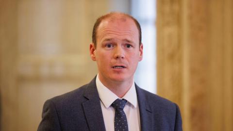 Gordon Lyons, with short brown hair, wearing a white shirt, navy tie and navy suit jacket.