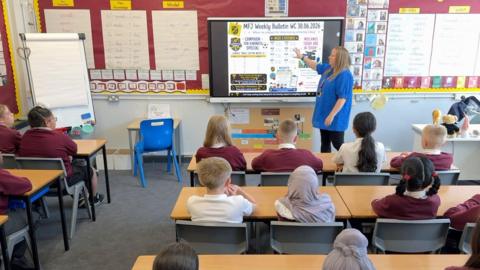 Schoolchildren sit behind desks in a classroom with a teacher stood at the front next to an interactive whiteboard.