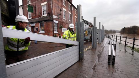 Two men wearing high-vis clothing and white hard hats slot metal beams into place, along the waterfront in Bewdley. The swollen River Severn can be seen on right, in front of the barrier, with a red-brick building behind the barrier that has white-framed windows, a green sign hanging on it, and decorative lights hanging from it.
