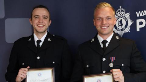 Two police officers holding certificates of commendation from the chief constable of Warwickshire Police. They are both wearing black police jackets, with a white shirt and black tie. The man on the left has short, dark hair and the man on the right has short, blonde hair with a quiff.