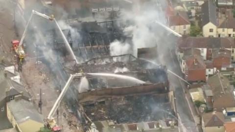 A burnt building with a collapsed roof is being hosed down by firefighters 