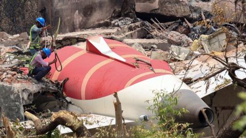 Two men wearing hard hats and shirts lower a rope at the crash site. The red and white plane is embedded into a building and surrounded by debris.