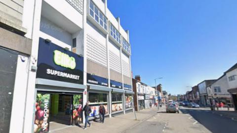 High Street in Scunthorpe taken from Google Streetview. There are people walking past a continental supermarket and cars driving down the street. It is a sunny day. 