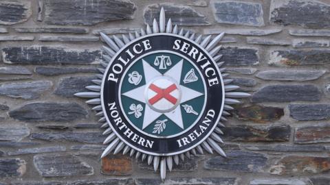 The PSNI crest mounted on a brick wall. It has the PSNI logo in the centre of a circular display with spikes coming out around the circumference.