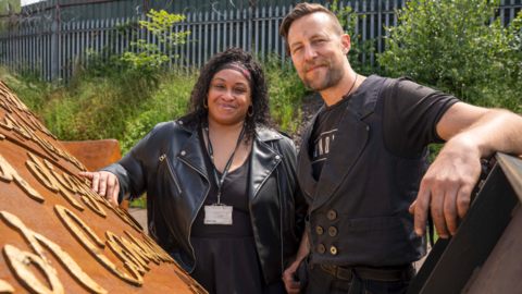 Victoria Murrain (left) and sculptor Luke Perry stand with the Windrush monument which is partially hidden from view in the park. What can be seen is a Corten steel colour with writing on it. The artists stand together behind it both wearing black and smiling at the camera. Victoria has long black hair and a scarf around her head. Luke has short brown hair and a beard.