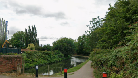 A pathway with the canal to the left and a large hedgerow to the right. The sky is cloudy and there are three black bollards at the start of the path. 