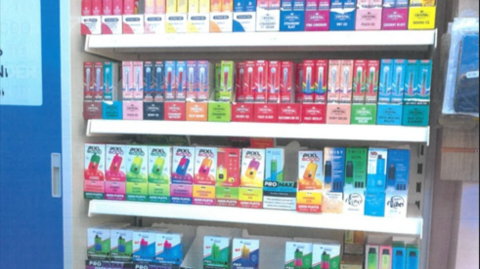 Different coloured vapes are positioned on several shelves in a shop next to a blue door. At least four shelves are visible.

