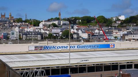 Photo of the roof of Guernsey harbour with a 'Welcome to Guernsey' sign. There are houses and buildings in the background and a blue sky.