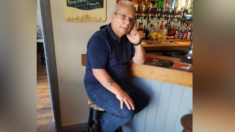A man in a blue t-shirt and jeans leaning on a bar in a pub. He is looking at the camera.