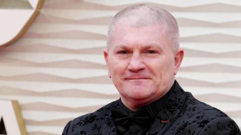 Ricky Hatton stands in front of a cream coloured wall smiling. He is wearing an embroidered black suit and tie. 