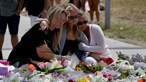 Three women with blonde hair look at floral tributes