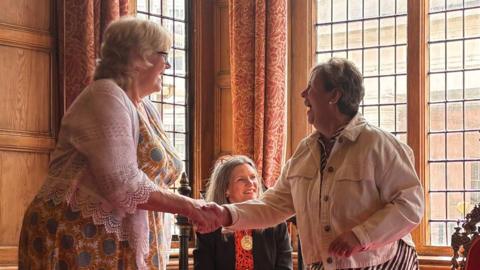 Anne Hope and Jenny Watkins shaking hands in an ornate, wood-panelled room. A woman in mayoral chains is sat in a chair behind them. The women are smiling.