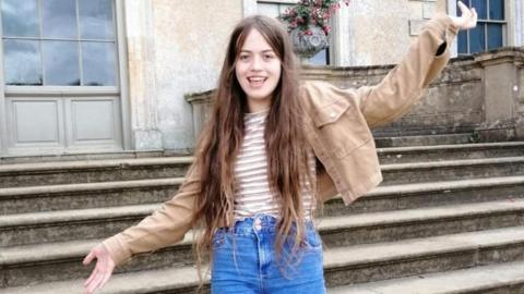 Image of Zara. She has brown hair and is wearing blue jeans and a light brown jacket. She is smiling for the camera
