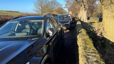 A row of cars parked on the roadside at Otley Chevin next to a wall.
