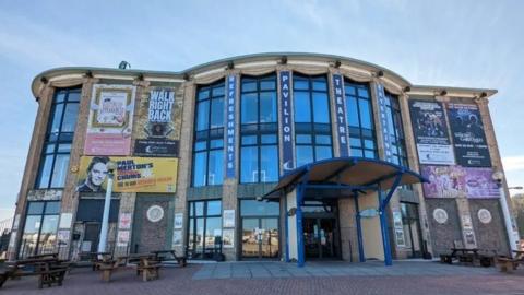 The front view of Weymouth Pavilion. It is a large building with a curved facade, which features large glass windows. There is a curved awning overlooking the front entrance, and large-scale posters of plays are affixed to the buildings two front-facing wings.