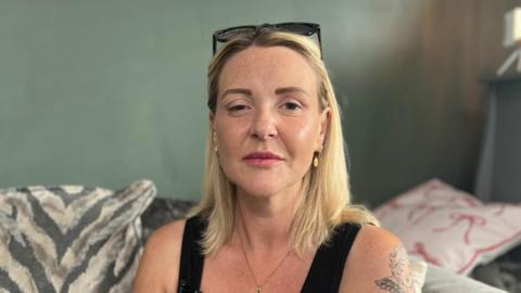 Nicola Fairly, a blonde woman in her 30s who is wearing a black vest and black sunglasses on her head. One side of her face is droopy which is a symptom of botulism. She has a tattoo on her left shoulder and is sitting on a sofa with cushions of different patterns.