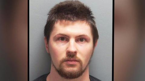 Custody image of 30-year-old Ethan Orchard. He has short dark hair, dark eyes, light complexion, and a light brown moustache and beard. He is wearing a black t-shirt.