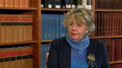 Lady Dorrian sits in a library, she has grey hair, wears a blue scarf and navy suit