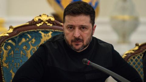 President of Ukraine Volodymyr Zelenskyy speaks at a meeting of the National Security and Defense Council on March 3, 2026 in Kyiv, Ukraine. He's leaning forward in his chair over a microphone, with a serious look on his face, and is dressed all in black