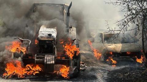 A burning tractor with smoke and flames rising from the charred frame that remains. There is also a car burning on the left of the tractor with ash and debris on the ground around the vehicles.