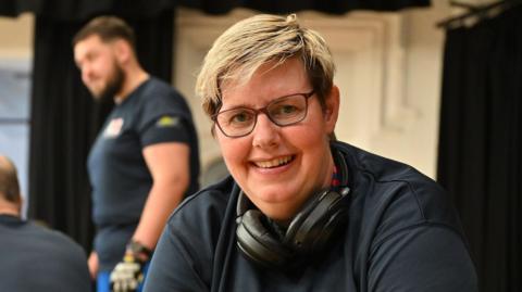 A woman is smiling at the camera and has headphones around her neck. 
She has very short blonde hair and glasses. She is wearing a blue top.
There is a man with a beard standing a few feet behind her talking to someone else.
