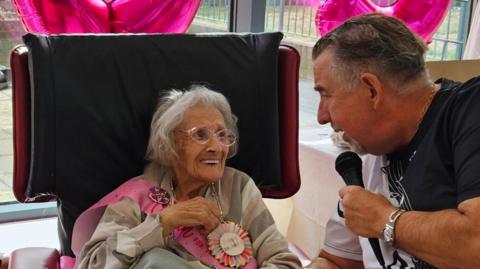 Ada Cook is on the left sat on a black and red chair. She has shoulder length grey hair and is wearing a pink birthday sash and a badge. On the right is a man wearing a white and black T-shirt holding a black microphone. 