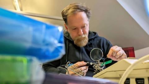 A man with a beard uses a soldering iron to fix a circuit board