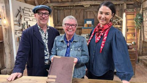 Chris Shaw, Gail Newsham, Sonnaz Nooranvary standing in the Repair Shop barn. Gail is in the middle holding the damaged scrapbook with is large with a red/brown cover wit patches of tape on it. She is wearing a denim jacket and is smiling, with short white hair and black-frames glasses