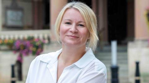 Sarah Ward, with long blonde hair, tied back, wearing a white shirt, looking at the camera and smiling, outside a building, which is blurred in the background, with flowers to the left. She has earrings in both ears. 