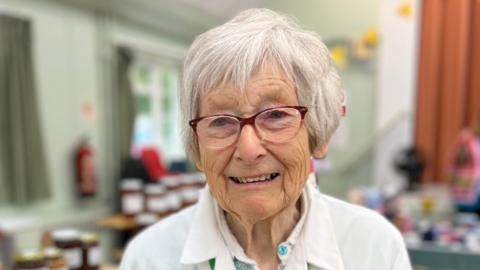 Dorothy Doel has short white hair and is wearing a white T-shirt. She is standing in a hall that has an indoor market where several items are on display across the room.