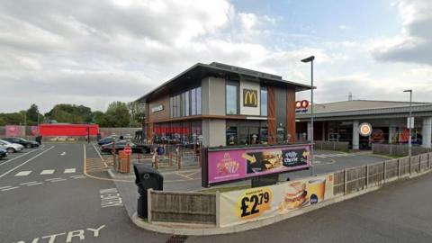 A two-storey branch of McDonald's with the golden arches on the front of the building and a drive-thru lane around it. A Tesco is visible behind it.