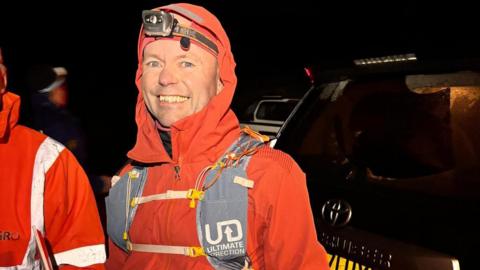 A man wrapped up in a red coat and wearing a head torch and a backpack, smiles. It is dark behind him. A car is visible on the right.