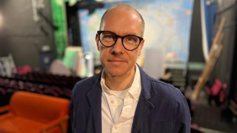 Derek Bond is standing in the middle of the picture with a blue jacket and white shirt - he has glasses and in the background is stage with theatrical props and furntiture.
