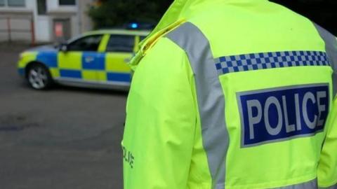 A person wearing a fluorescent police jacket with the word police written across the back. A police car is in the foreground with its blue lights on