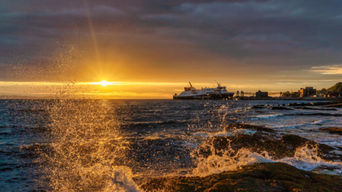 Water splashing against rocks on Arran. The sky is grey and orange. The water in the foreground is white and is leaping up on the rocks. In the background is a ferry which is dark and white.