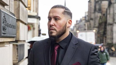 A man with short brown hair and a beard outside a court building. He is wearing a grey suit jacket, black shirt and maroon tie