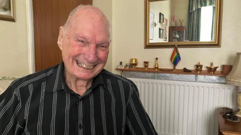 A picture of an elderly man wearing a black shirt with gray stripes on it. There is a mirror in the background and a pride flag.